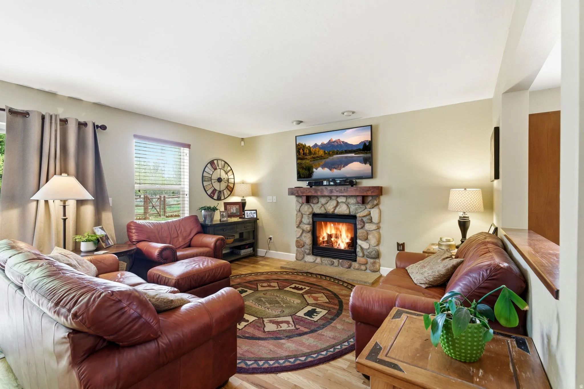 Living room featuring a stone-clad fireplace with a wood mantel, wood-finish flooring, and a large window with blinds