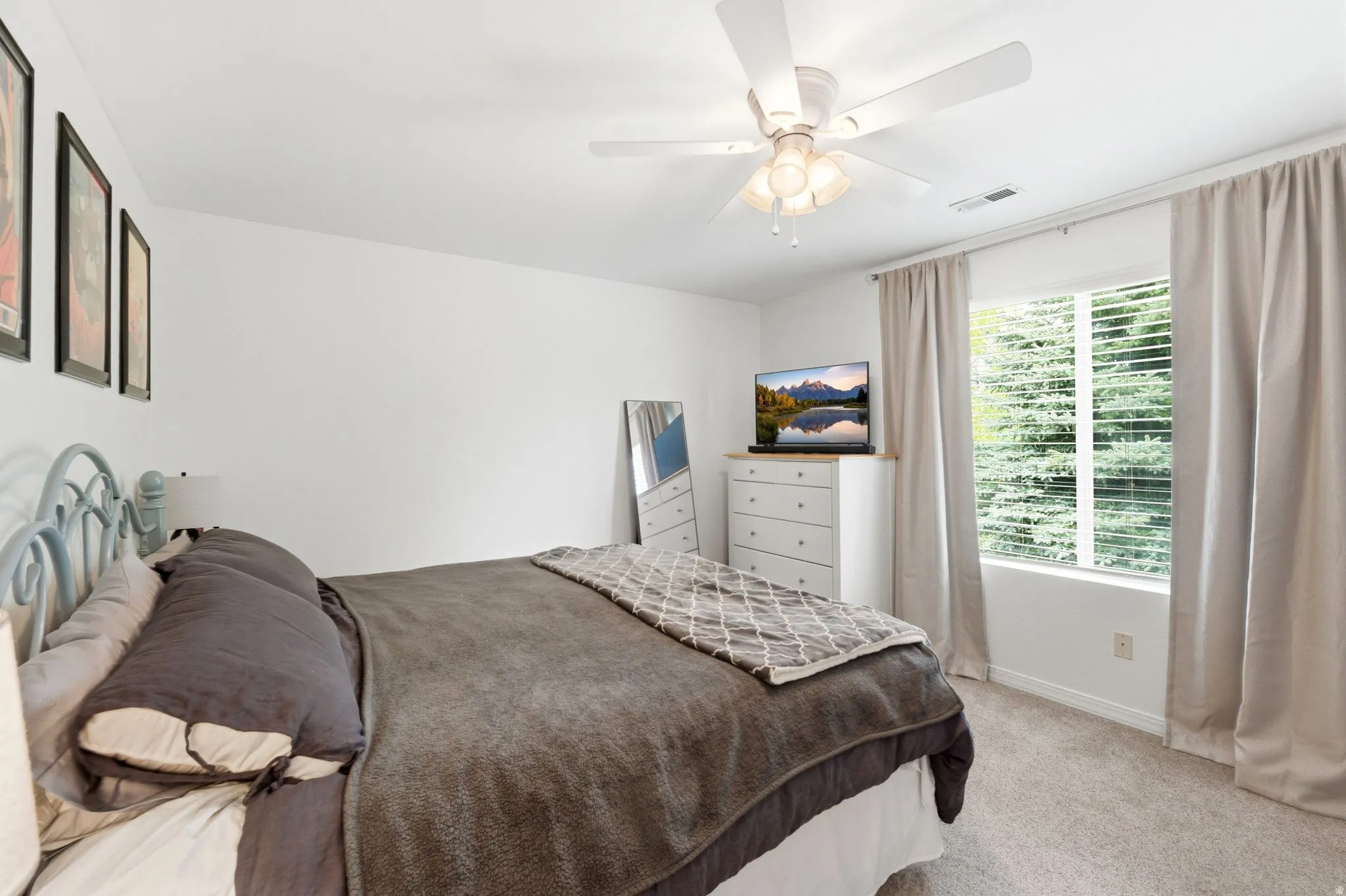 Carpeted bedroom featuring a ceiling fan, a window with horizontal blinds, and neutral-toned curtains