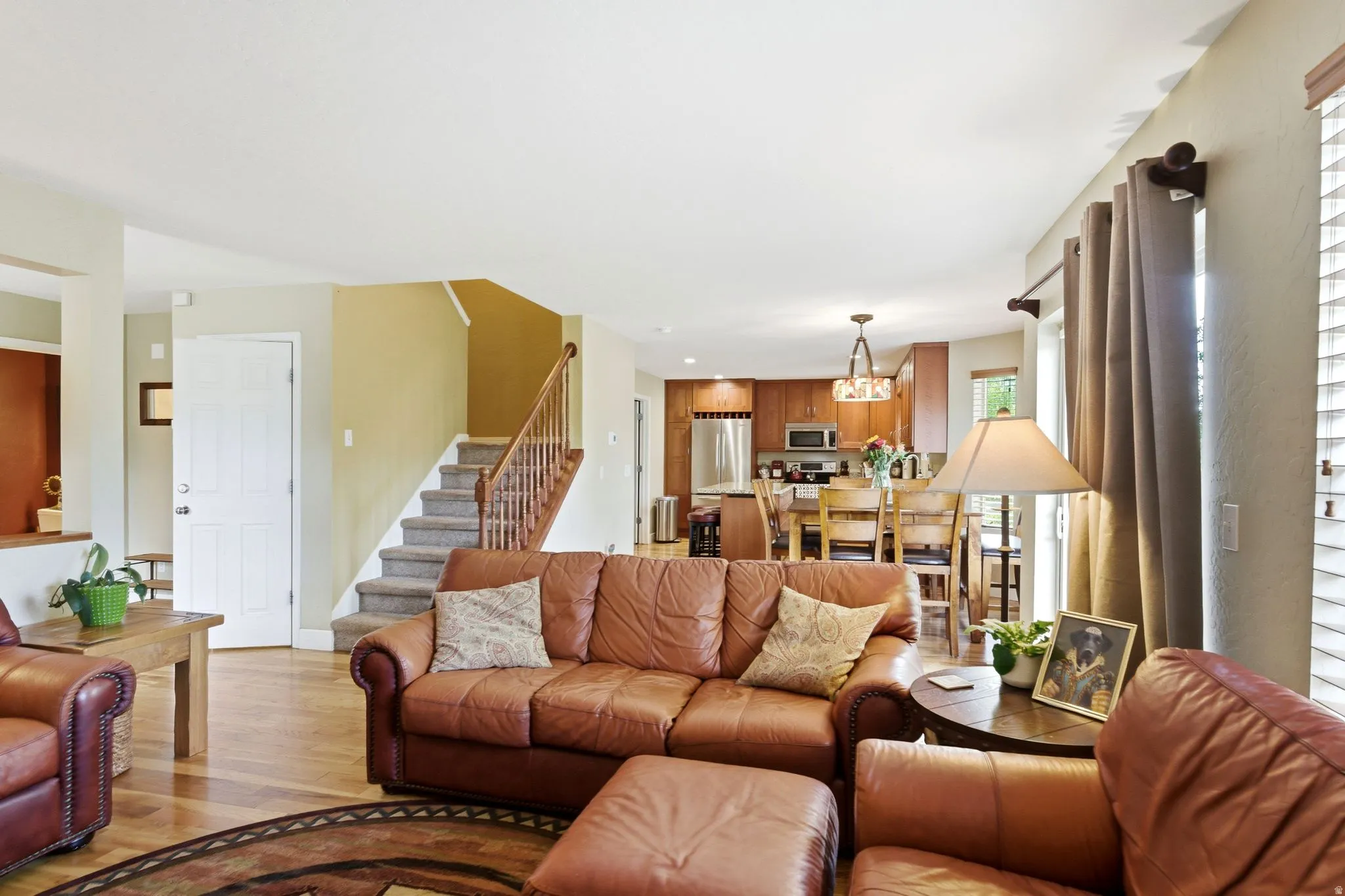 Open-concept living space featuring wood-finish flooring, a carpeted staircase with wood banister, and a kitchen with wood cabinetry, stainless steel appliances, and a central island