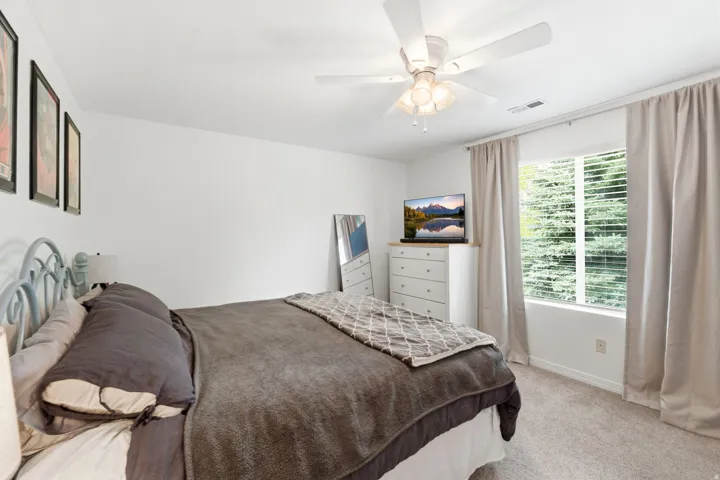 Carpeted bedroom featuring a ceiling fan, a window with horizontal blinds, and neutral-toned curtains