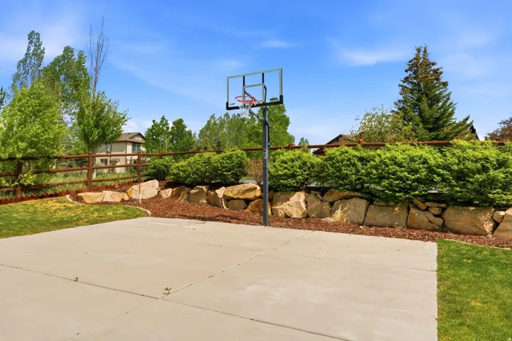 Concrete sport court with a portable basketball hoop, bordered by a split-rail fence and mature landscaping