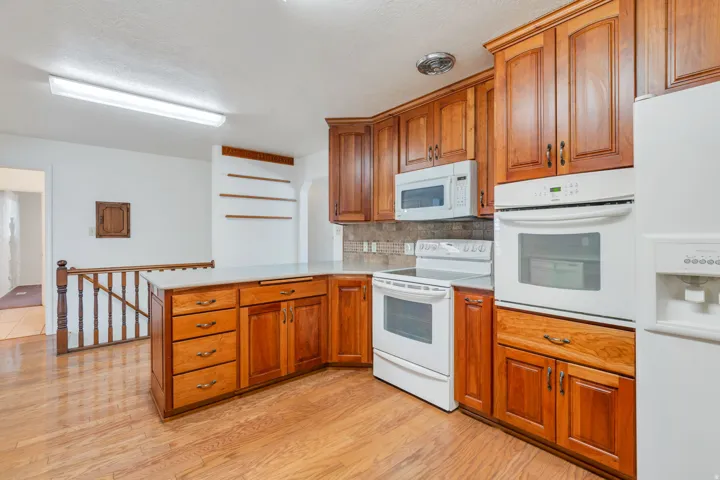 Kitchen featuring wood-finish flooring, wood cabinetry, white appliances, a tile backsplash, and a breakfast bar