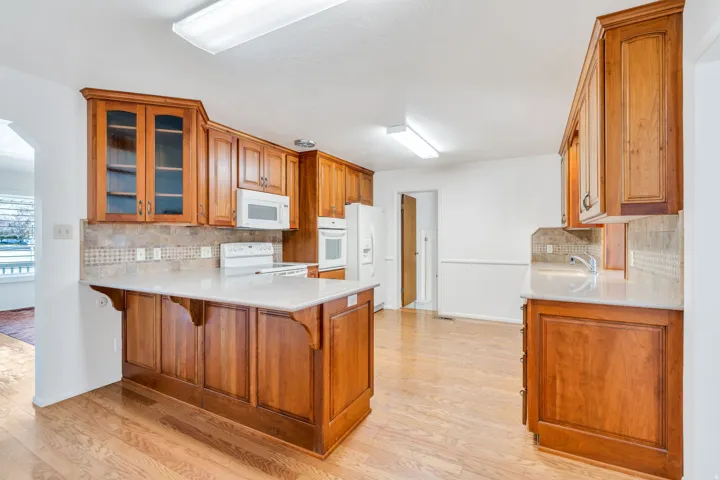 Kitchen featuring wood cabinetry, white appliances, light countertops, wood-finish flooring, and a tiled backsplash