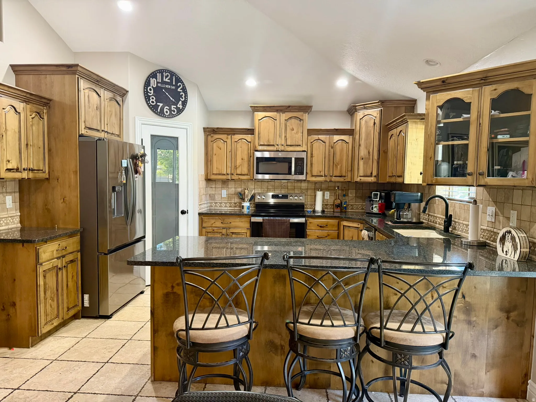 Spacious kitchen featuring a large central island with granite countertops, wood-finish cabinetry, stainless steel appliances, tile flooring, and vaulted ceilings. Refirgerator included.