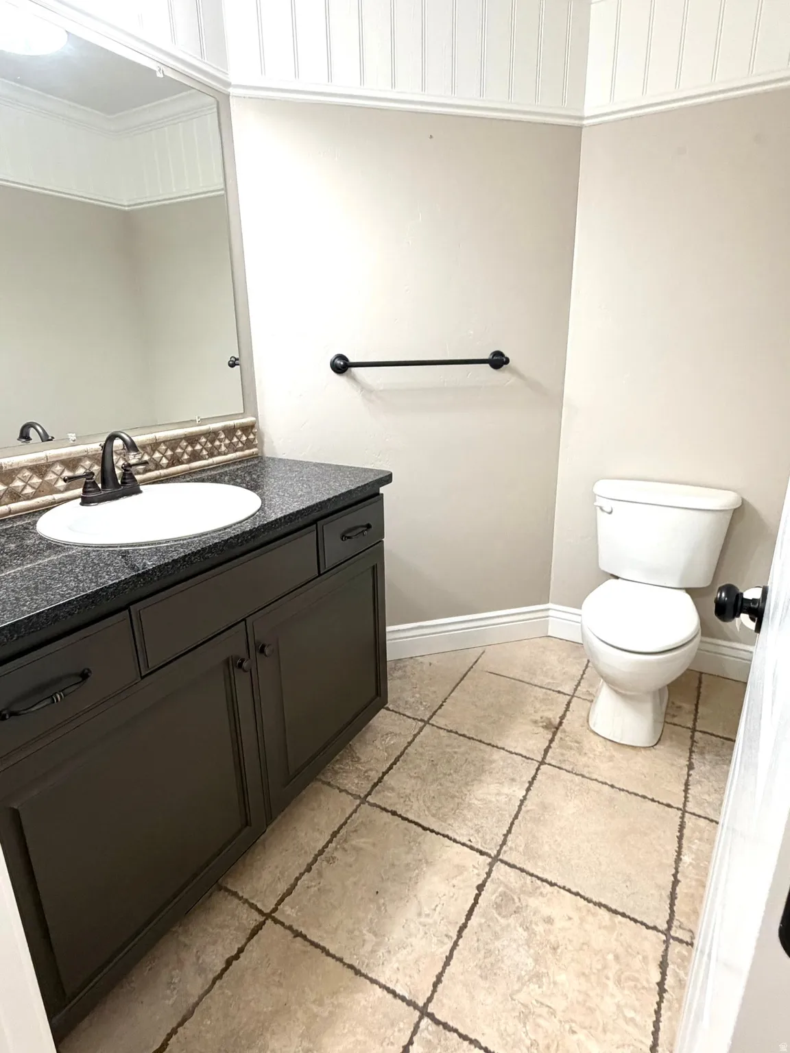 Half bath featuring a dark vanity with a white basin sink and dark granite countertop, a decorative travertine tiled backsplash, a white toilet, and tiled flooring