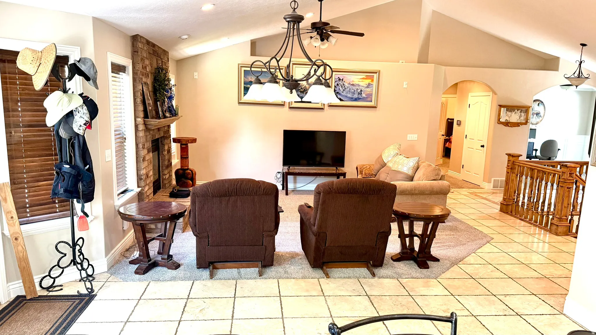 Vaulted ceilings with a ceiling fan and chandelier, stone-faced fireplace with a wood mantle, broad arched entryway, wood-finish railing, and light-toned tile flooring