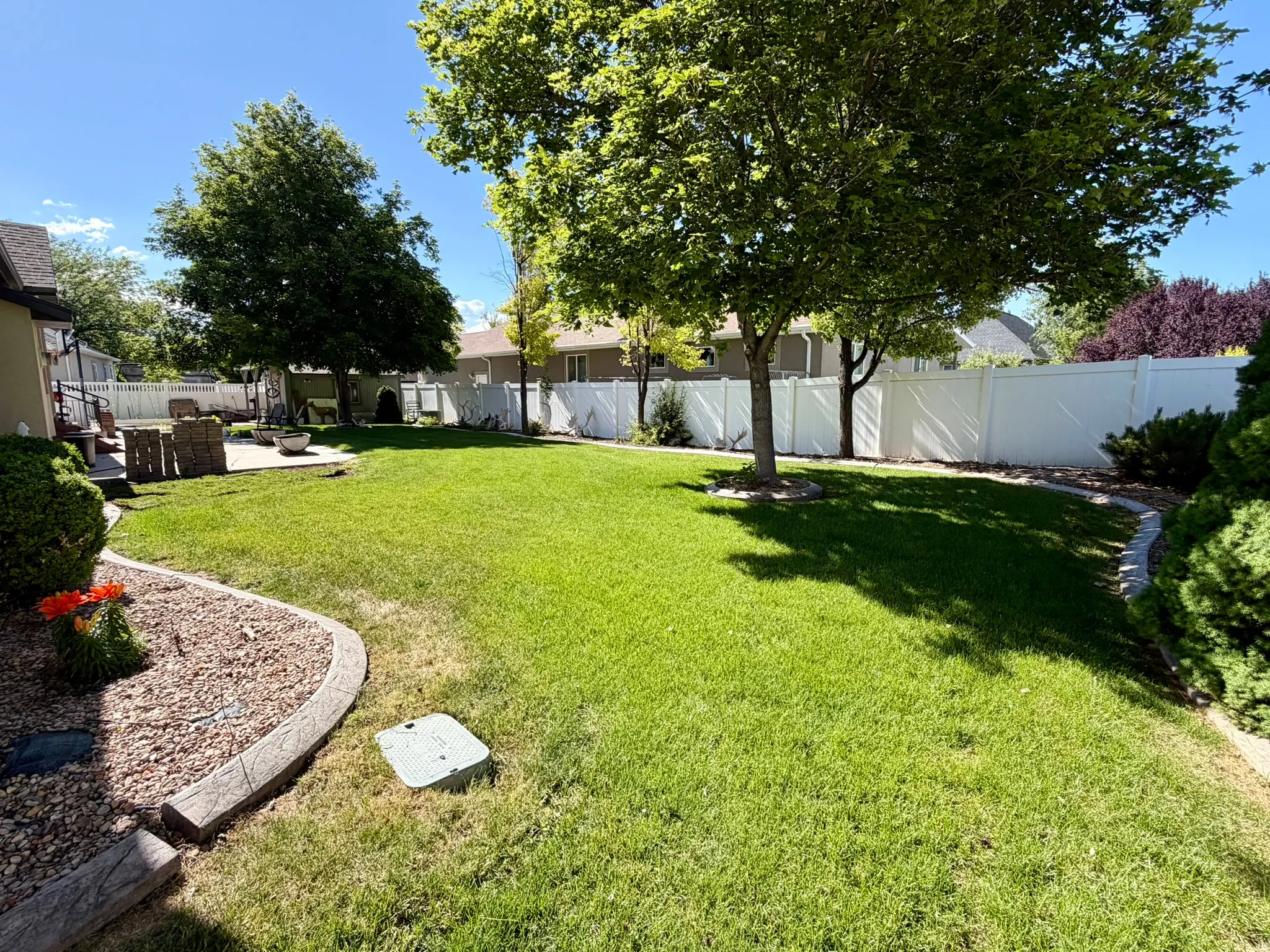 Expansive backyard featuring a lush lawn, mature trees, concrete patio, white privacy fencing, and decorative rock landscaping with concrete curbing