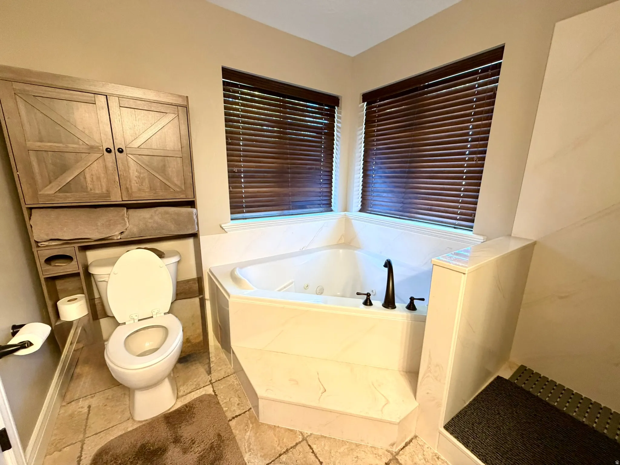 Primary corner soaking tub with dark bronze fixtures, framed by two windows with dark wood blinds, set on light-colored tile flooring