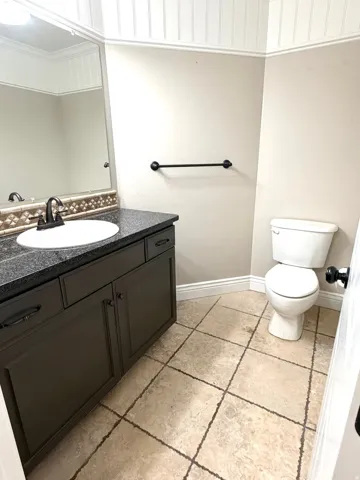 Half bath featuring a dark vanity with a white basin sink and dark granite countertop, a decorative travertine tiled backsplash, a white toilet, and tiled flooring