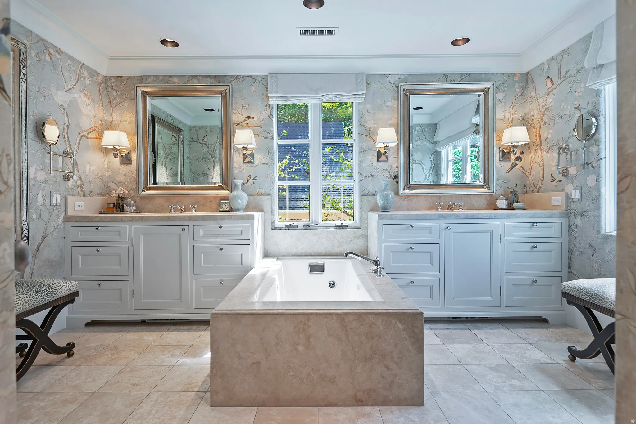 Spacious bathroom featuring a built-in soaking tub with stone surround, dual vanities with integrated sinks, framed mirrors, and decorative wall sconces