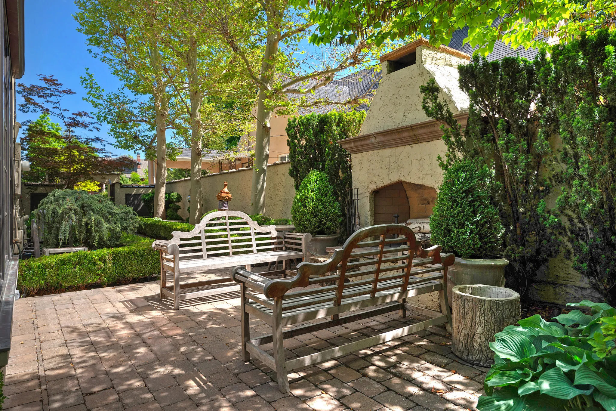 Brick paver patio featuring an outdoor fireplace, manicured hedges, and mature trees providing natural shade