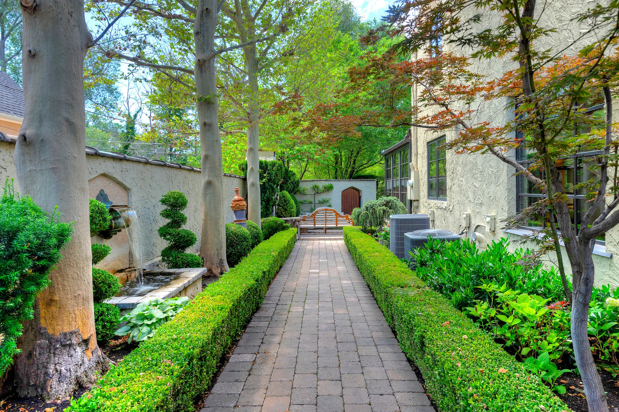 Manicured garden path featuring brick pavers, formal hedges, mature trees, a built-in fountain, and stucco exterior with dark-framed windows