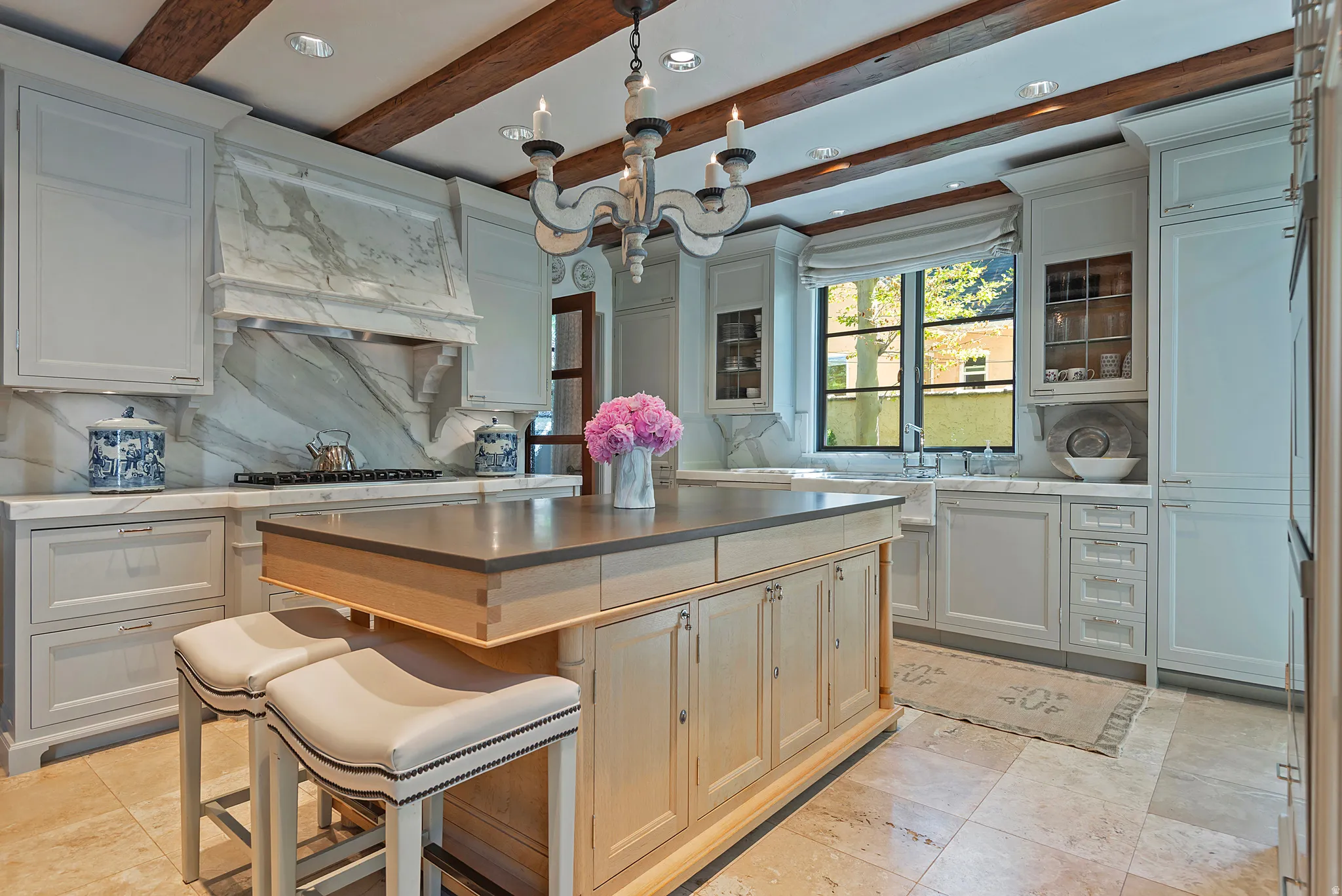Kitchen featuring a substantial central island with integrated seating, exposed wood ceiling beams, light-toned custom cabinetry, a statement range hood with a marble backsplash, and a large window above the farmhouse-style sink