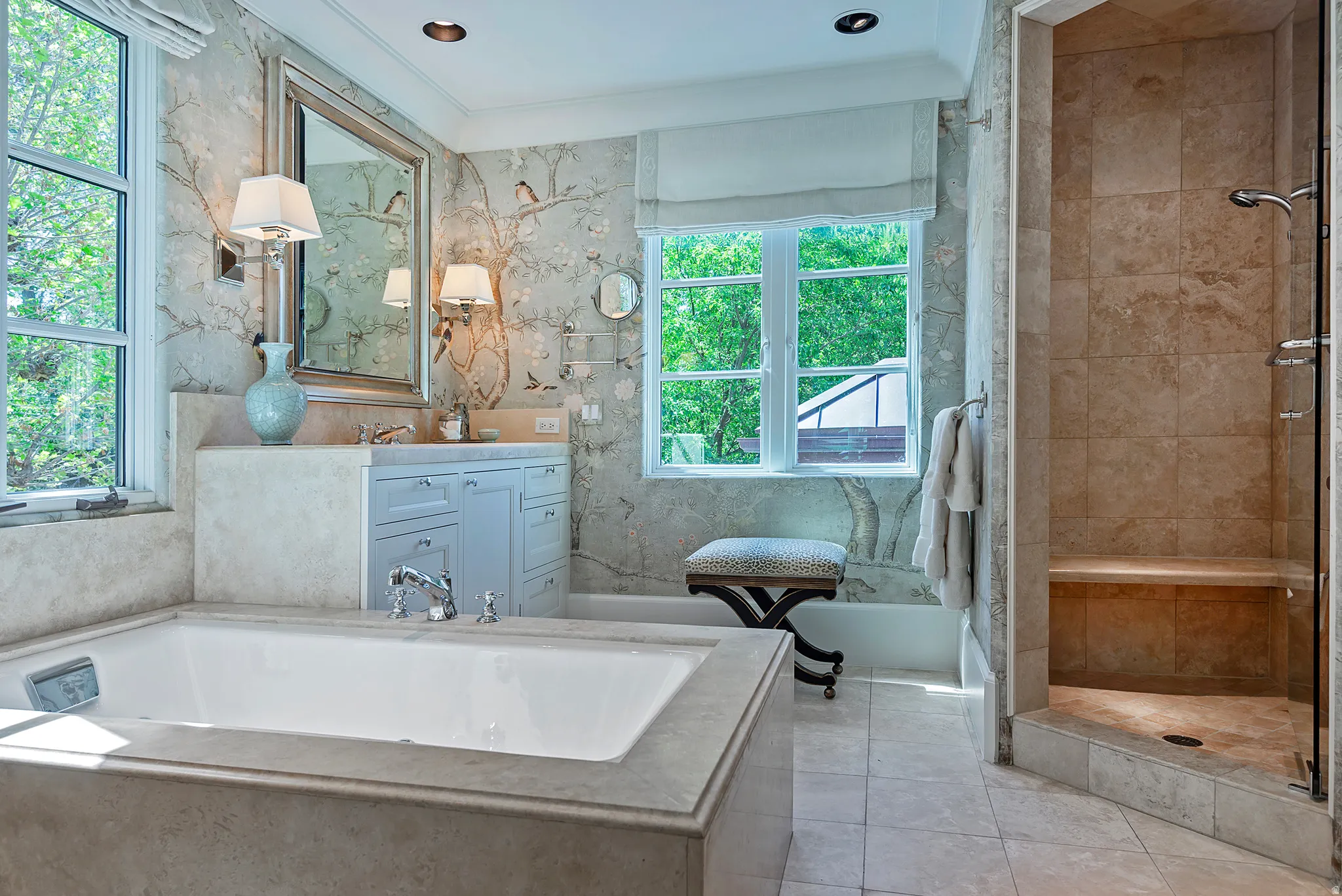 Spacious bathroom featuring a built-in soaking tub, separate tiled shower with a bench, dual windows, a vanity with under-sink cabinetry, and wall-mounted sconces