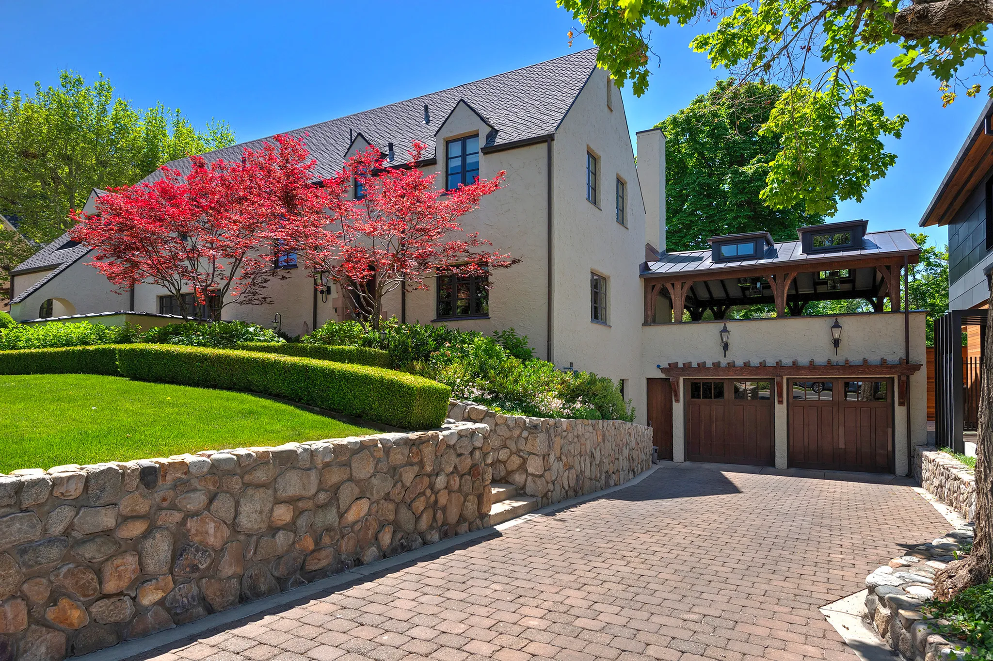 Stucco exterior residence with a shingled roof, featuring a paver driveway, stone retaining walls, and manicured landscaping
