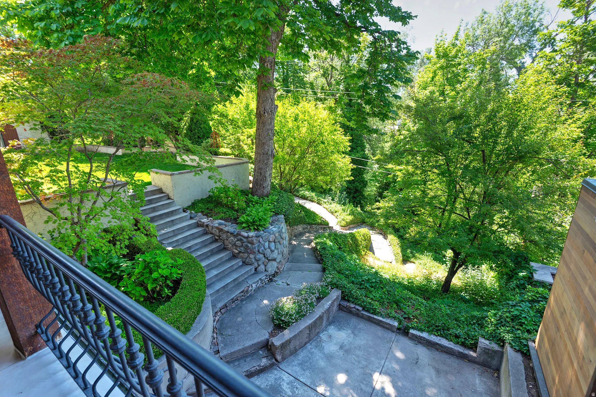 Tiered landscaping featuring concrete steps, natural stone retaining walls, mature trees, manicured hedges, and lush ground cover