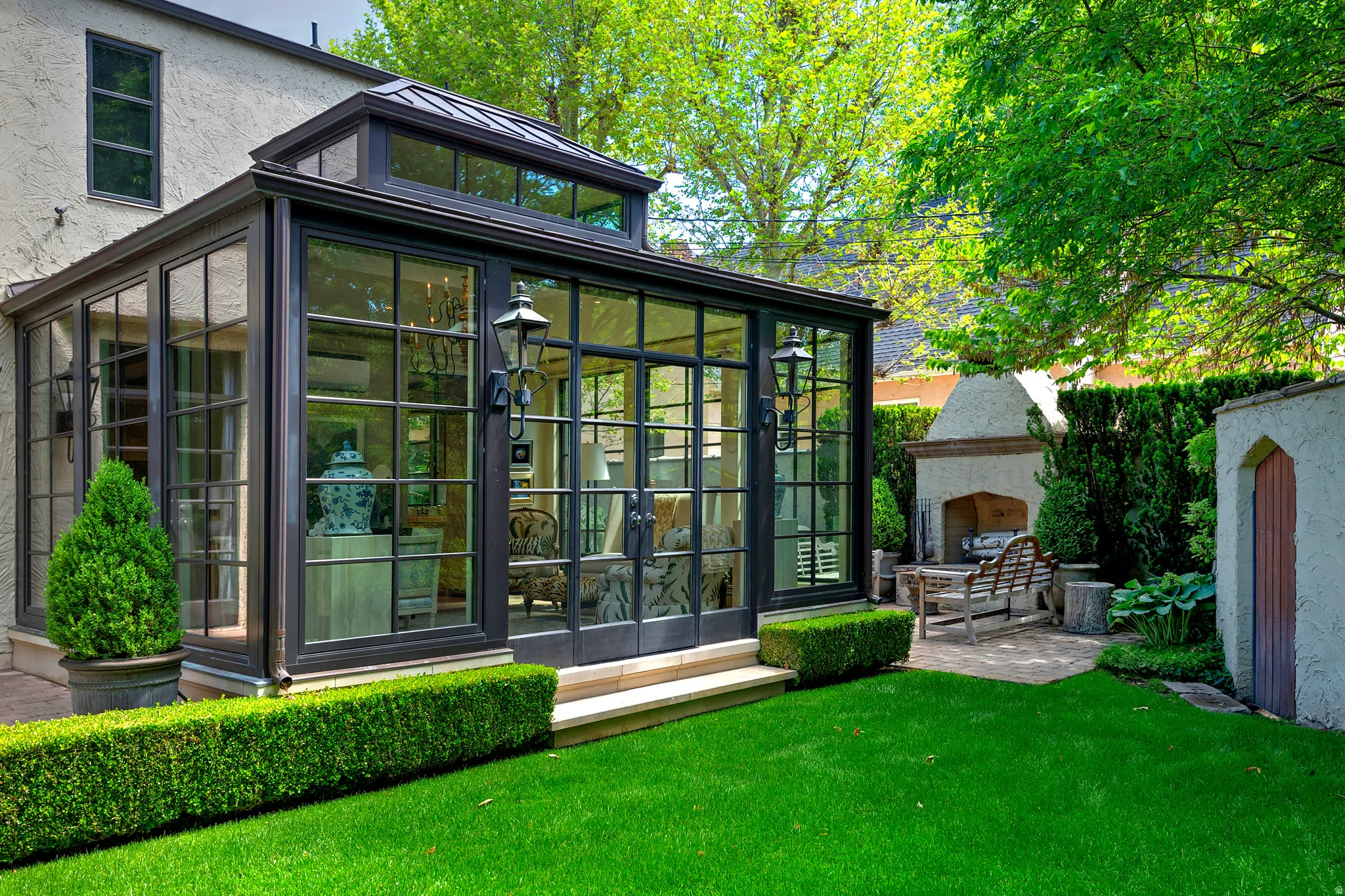 Sunroom addition with black-framed windows and a glass roof, featuring double doors opening to a paved patio with an outdoor fireplace