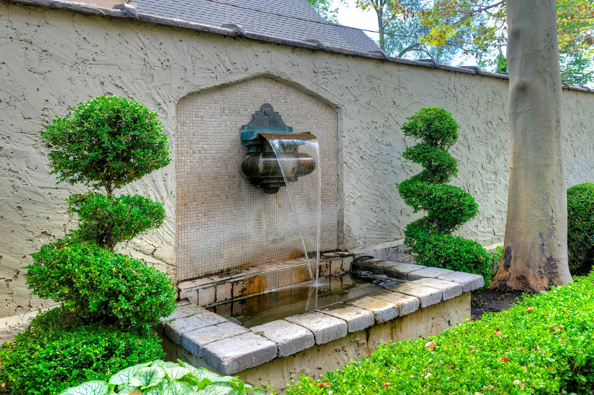 Stone-edged water feature with a mosaic tile backdrop, flanked by manicured topiary shrubs and a textured stucco wall