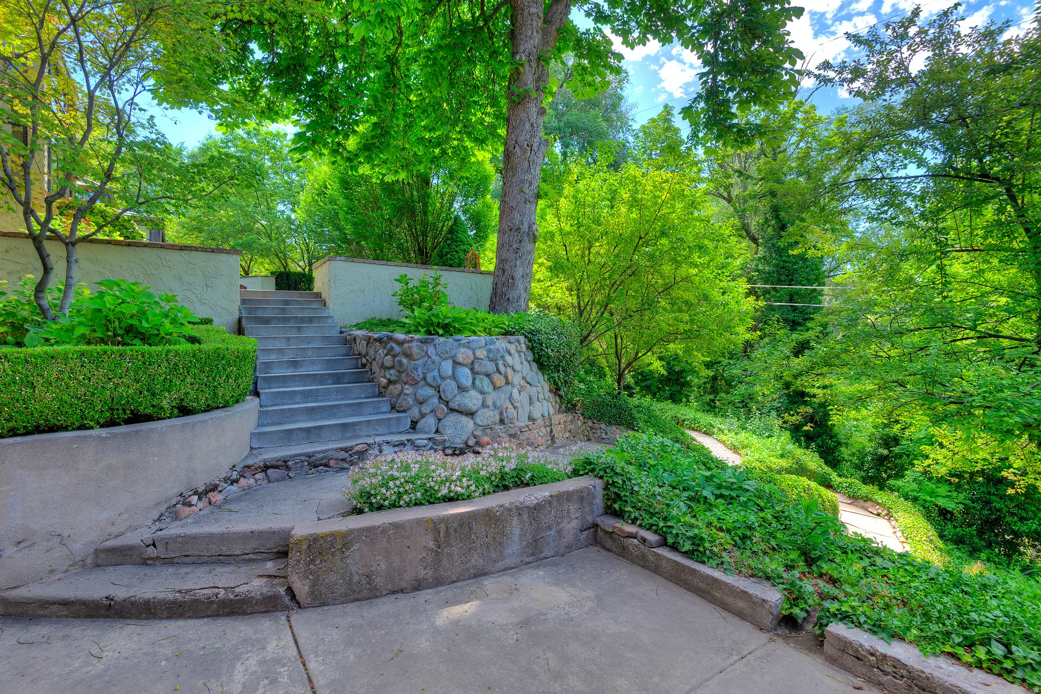 Terraced landscaping featuring concrete steps, stucco retaining walls, stone accents, mature trees, and established plantings
