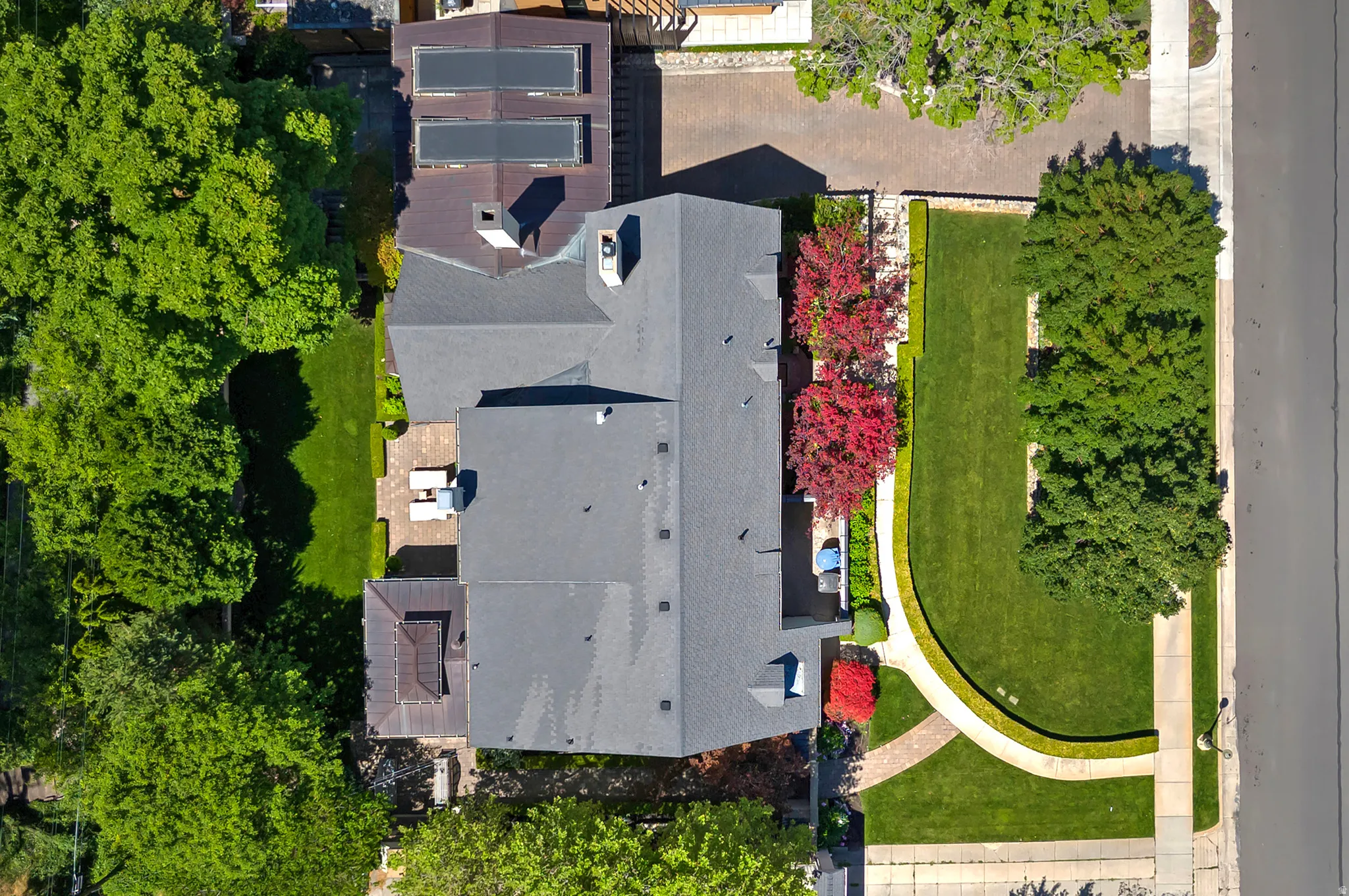 Expansive property featuring a grey shingle roof, mature green trees, vibrant red foliage, a meticulously maintained lawn, and a light-colored paved patio