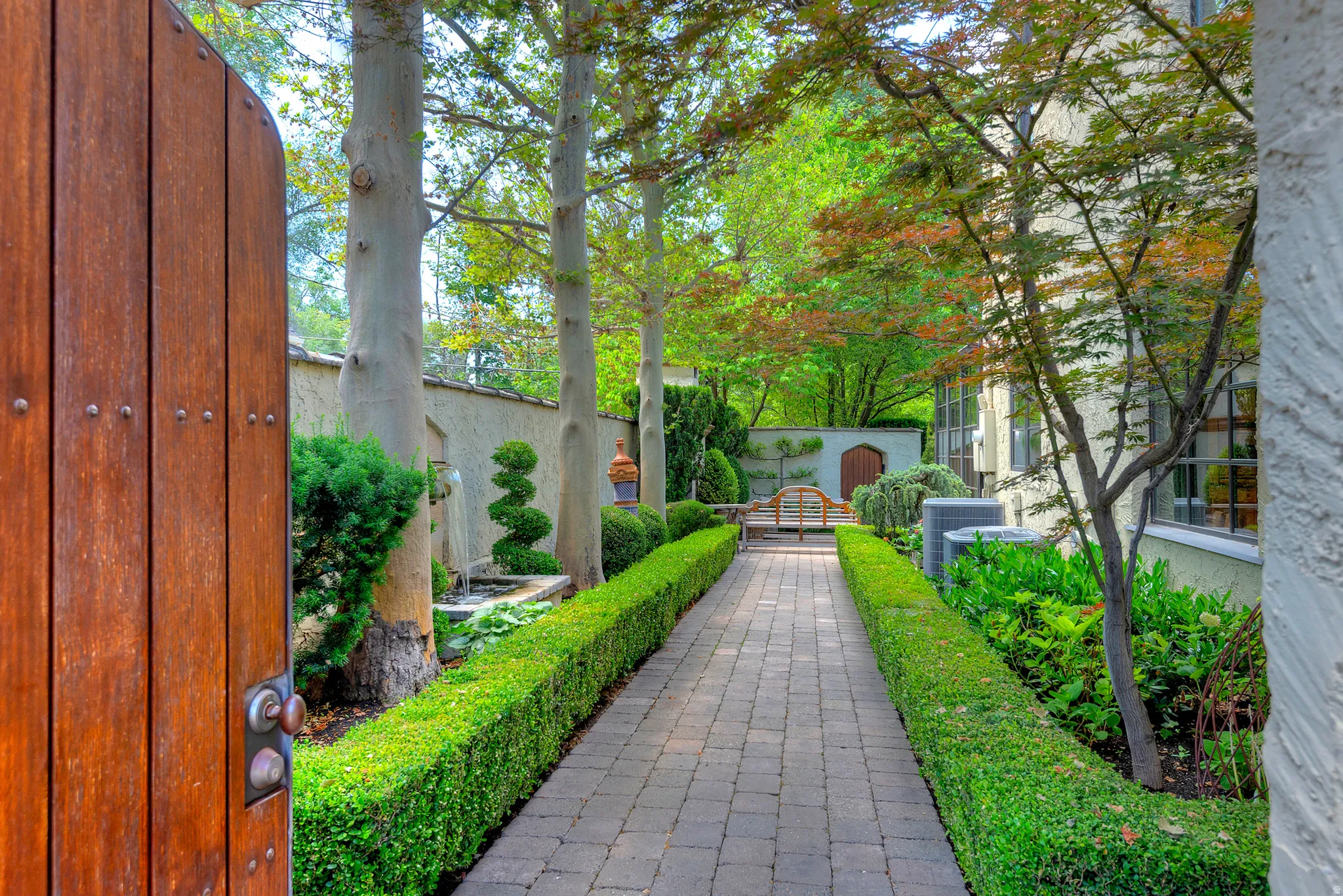 Private garden entry featuring a solid wood gate with iron hardware, a paver pathway, manicured hedges, mature trees, and a decorative garden wall with integrated seating