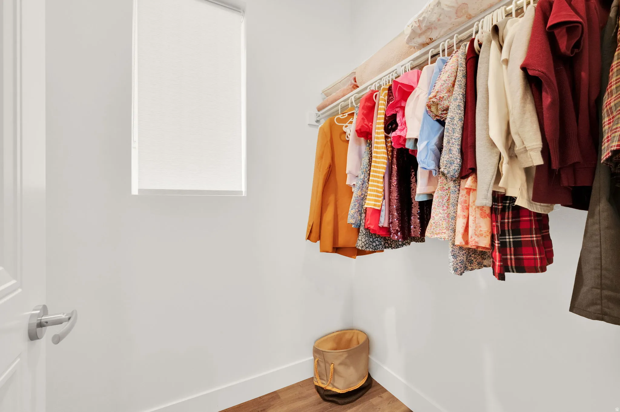 Walk-in closet featuring wood-finish flooring, white walls, a single window with roller shade, and built-in clothes hanging rods with an overhead shelf