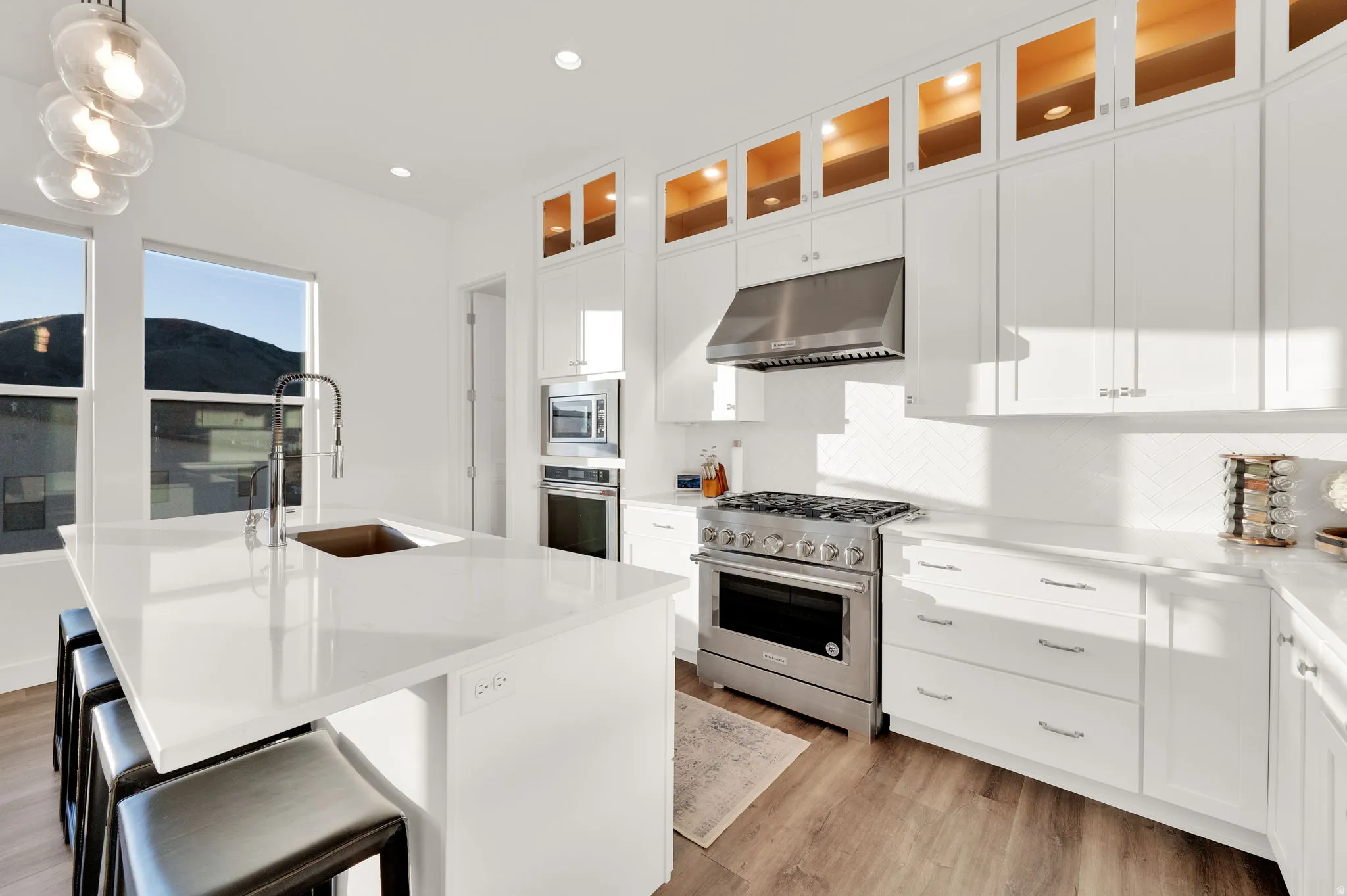Bright kitchen featuring an expansive central island with an undermount sink, stainless steel appliances, white cabinetry with upper glass-front illuminated cabinets, and wood-finish flooring