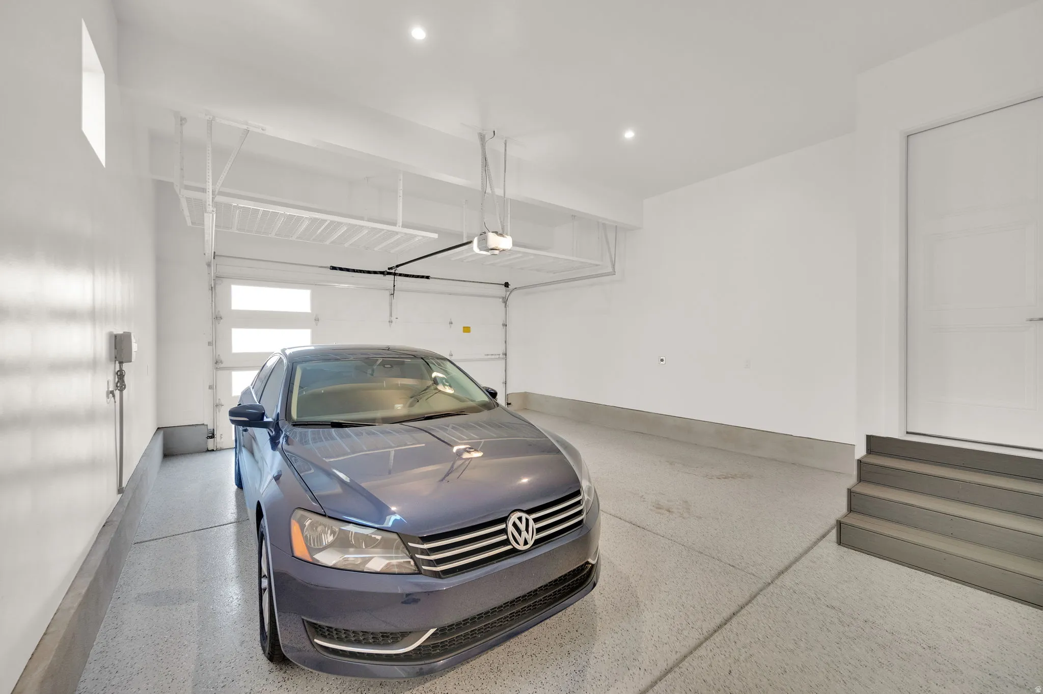 Spacious garage featuring a durable epoxy floor, an automatic overhead door with frosted glass panels, recessed ceiling lighting, a dedicated entry door, and a three-step staircase