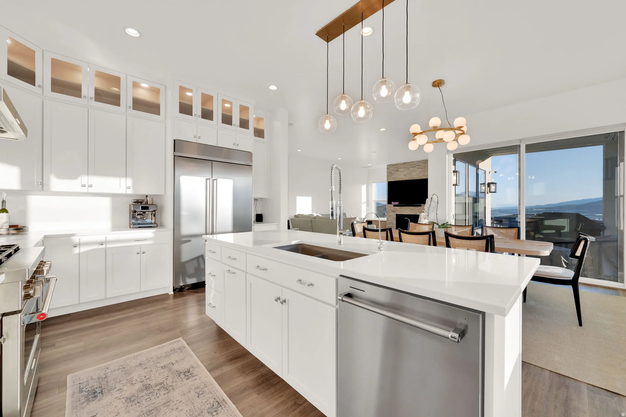 Gourmet kitchen featuring a large central island with an undermount sink, stainless steel appliances, white cabinetry with upper glass-front display cabinets, and wood-finish flooring