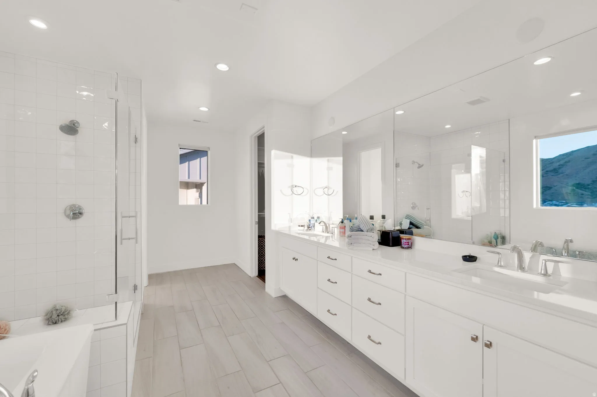 Bright bathroom featuring a dual vanity, expansive mirror, wood-finish flooring, and a glass-enclosed shower with a separate tub