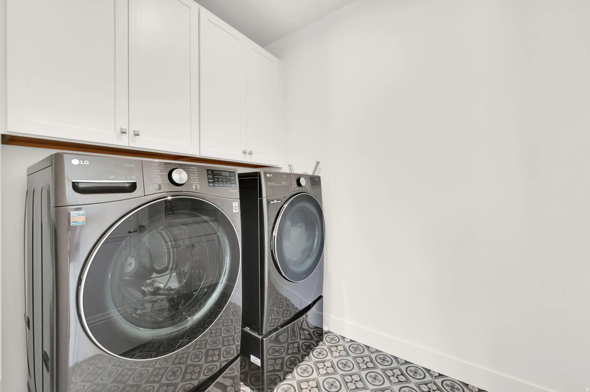 Laundry area featuring patterned floor tiles, white walls, and upper cabinetry with silver hardware