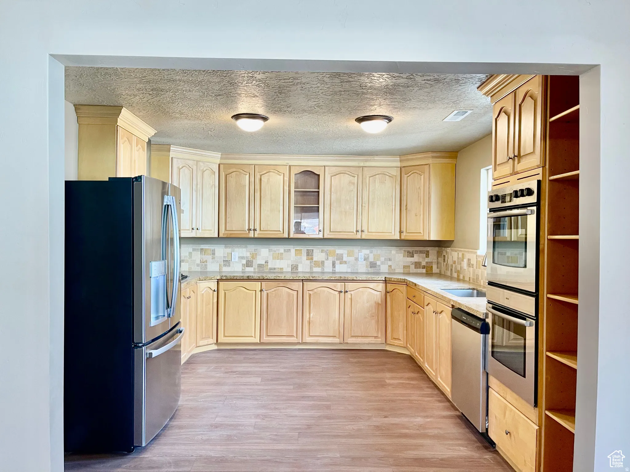 Kitchen featuring extensive light wood cabinetry, stone-finish countertops, tile backsplash, stainless steel appliances, and wood-finish flooring