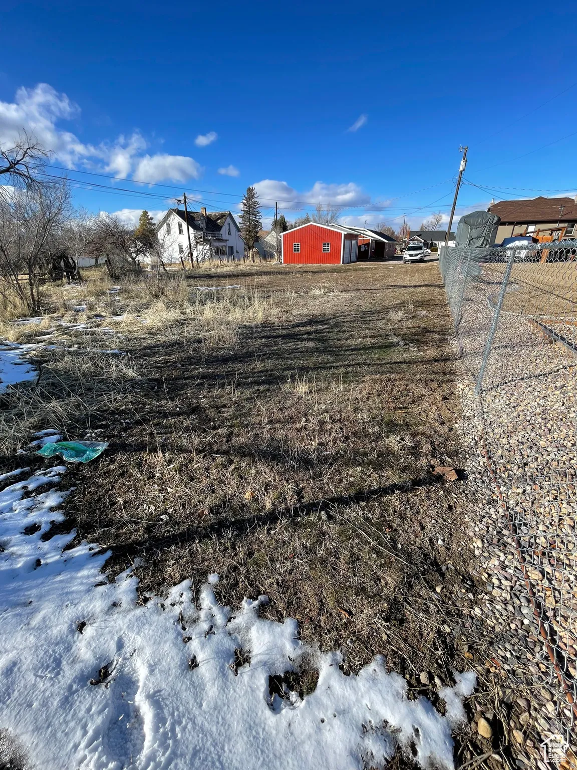 Expansive open lot featuring a red accessory building, partially visible chain-link fencing, gravel border, and a clear blue sky