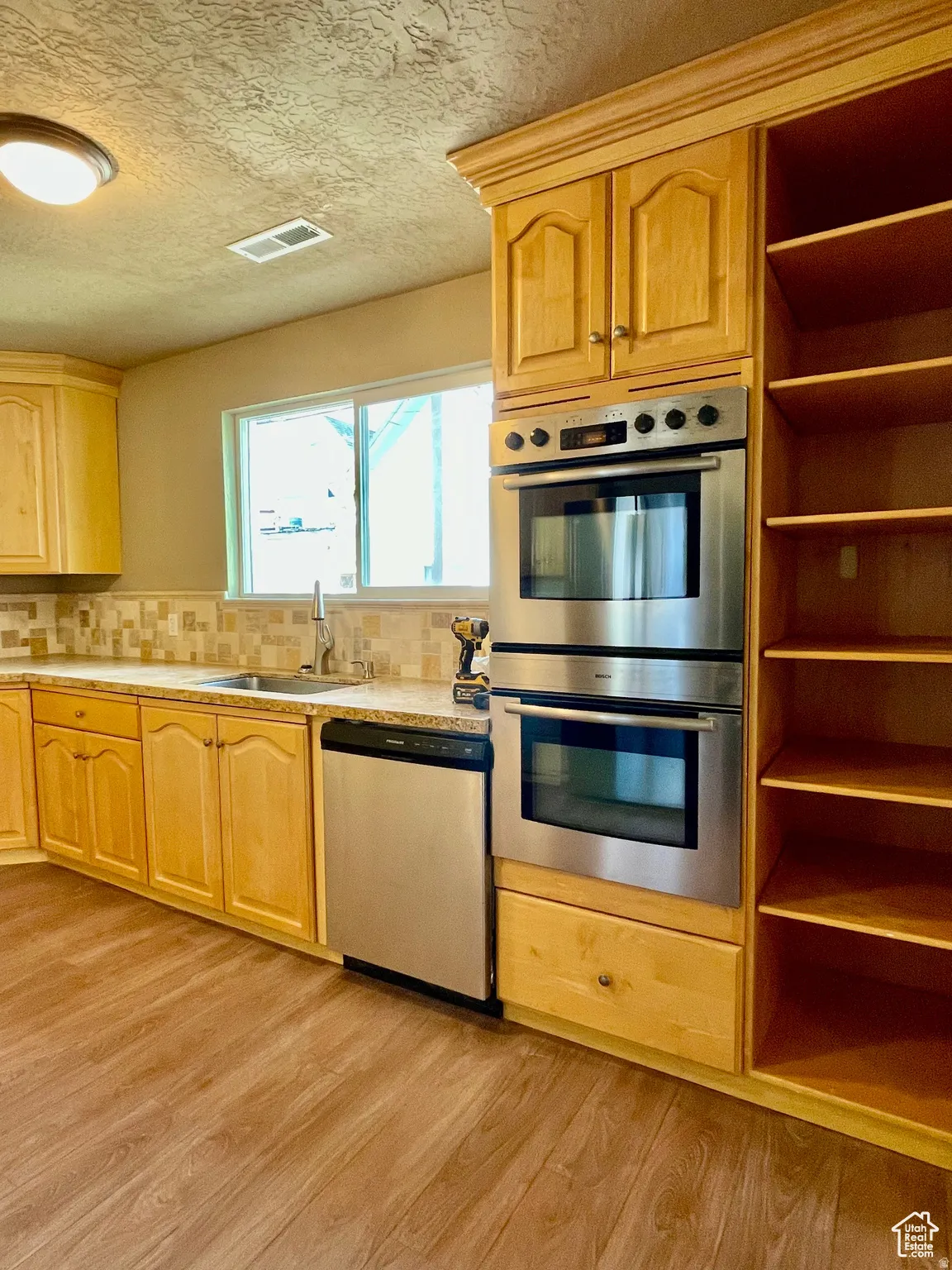Kitchen featuring light wood cabinetry, stainless steel double wall ovens, a coordinating dishwasher, wood-finish flooring, and a patterned tile backsplash