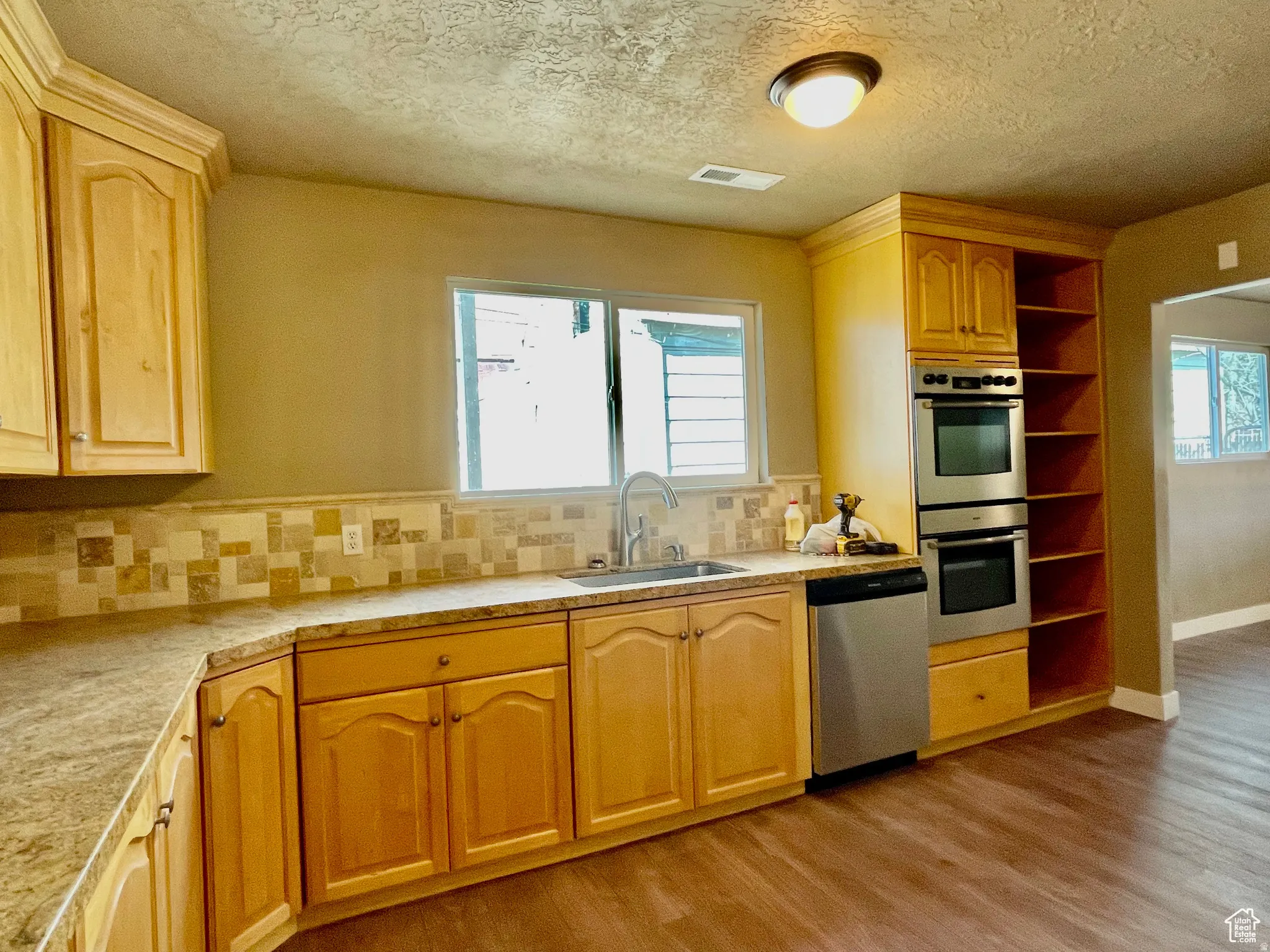 Kitchen featuring light wood-finish cabinetry, a stainless steel double wall oven, a stainless steel dishwasher, wood-finish flooring, and a tiled backsplash