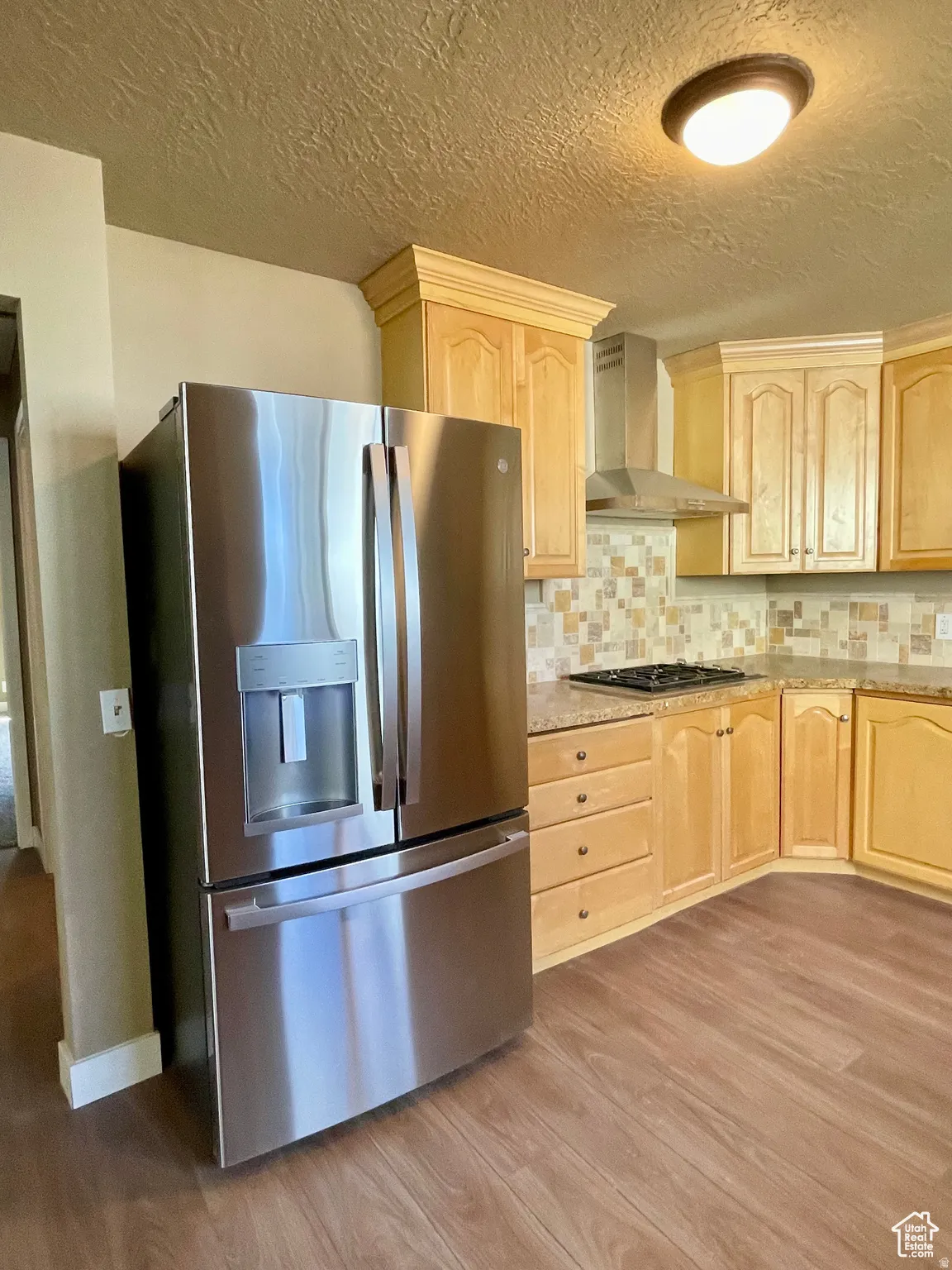 Stainless steel French door refrigerator, wood-finish flooring, light wood cabinetry, stone-look countertops, and a tiled backsplash