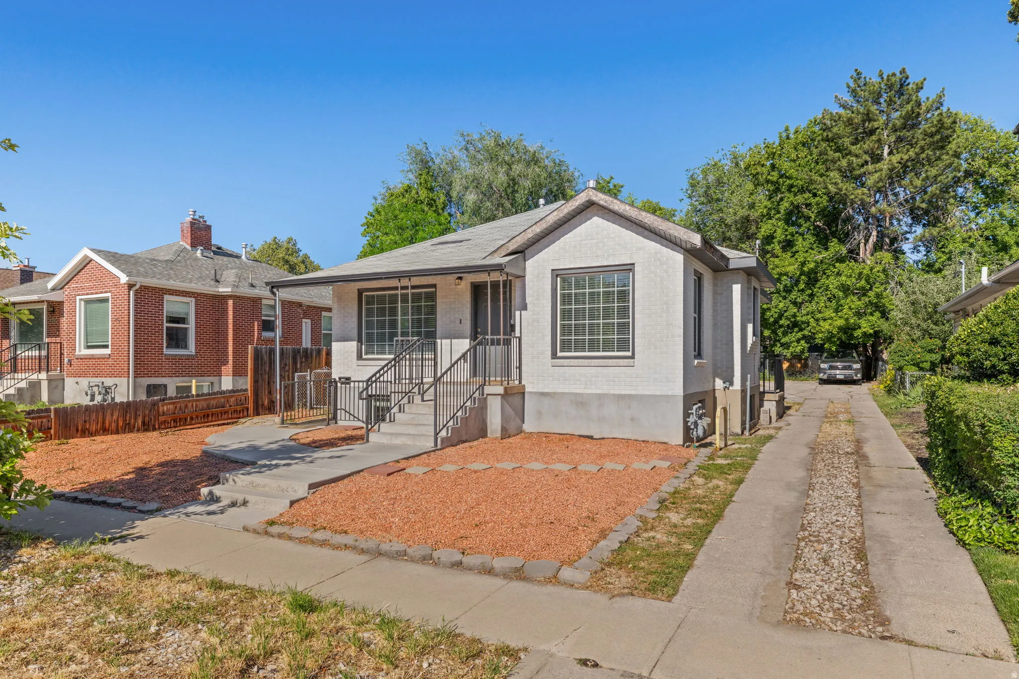 Stucco exterior with a peaked roofline, front porch with black metal railings, a dedicated concrete driveway, and an established tree-lined perimeter