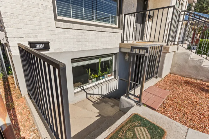 Exterior entry featuring concrete stairs, black metal railings, and a window well with a security grate