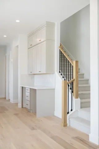 Built-in desk area with white cabinetry, light-toned wood-finish flooring, and a carpeted staircase featuring a light wood handrail and black metal balusters