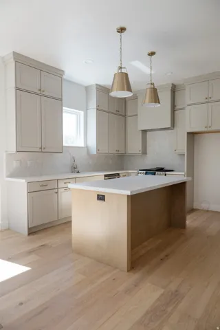 Kitchen featuring light-finish wood flooring, a central island with a light-toned countertop, perimeter cabinetry with light-toned countertops and backsplash, and two brass pendant lights