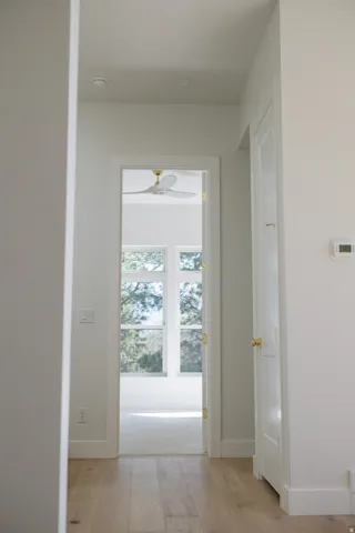 Hallway featuring light wood-finish flooring, white trim, and a doorway opening to a room with large windows, light carpeting, and a ceiling fan