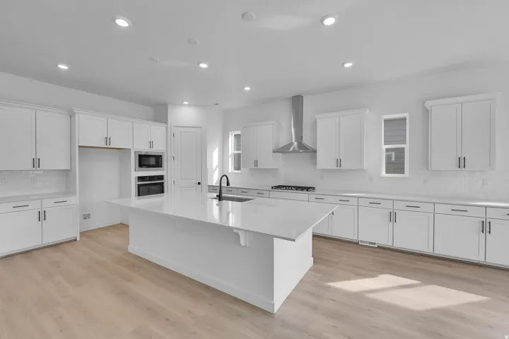 Spacious kitchen featuring an expansive island with a white countertop, extensive white cabinetry with dark hardware, wood-finish flooring, recessed lighting, and a stainless steel range hood