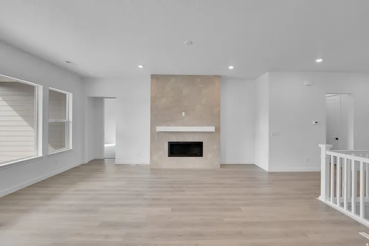 Spacious living area featuring light wood-finish flooring, a contemporary fireplace with a light-toned surround and white mantel, multiple windows, and recessed lighting