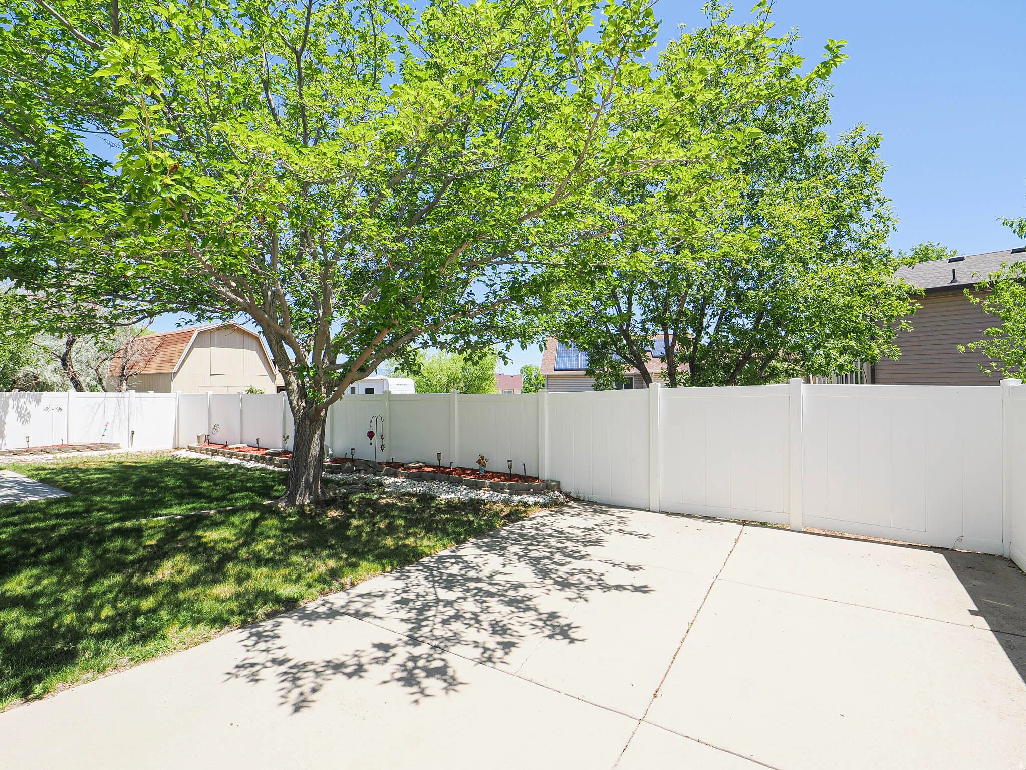 Private backyard with concrete patio, mature tree, manicured lawn, white vinyl fencing, and decorative rock landscaping
