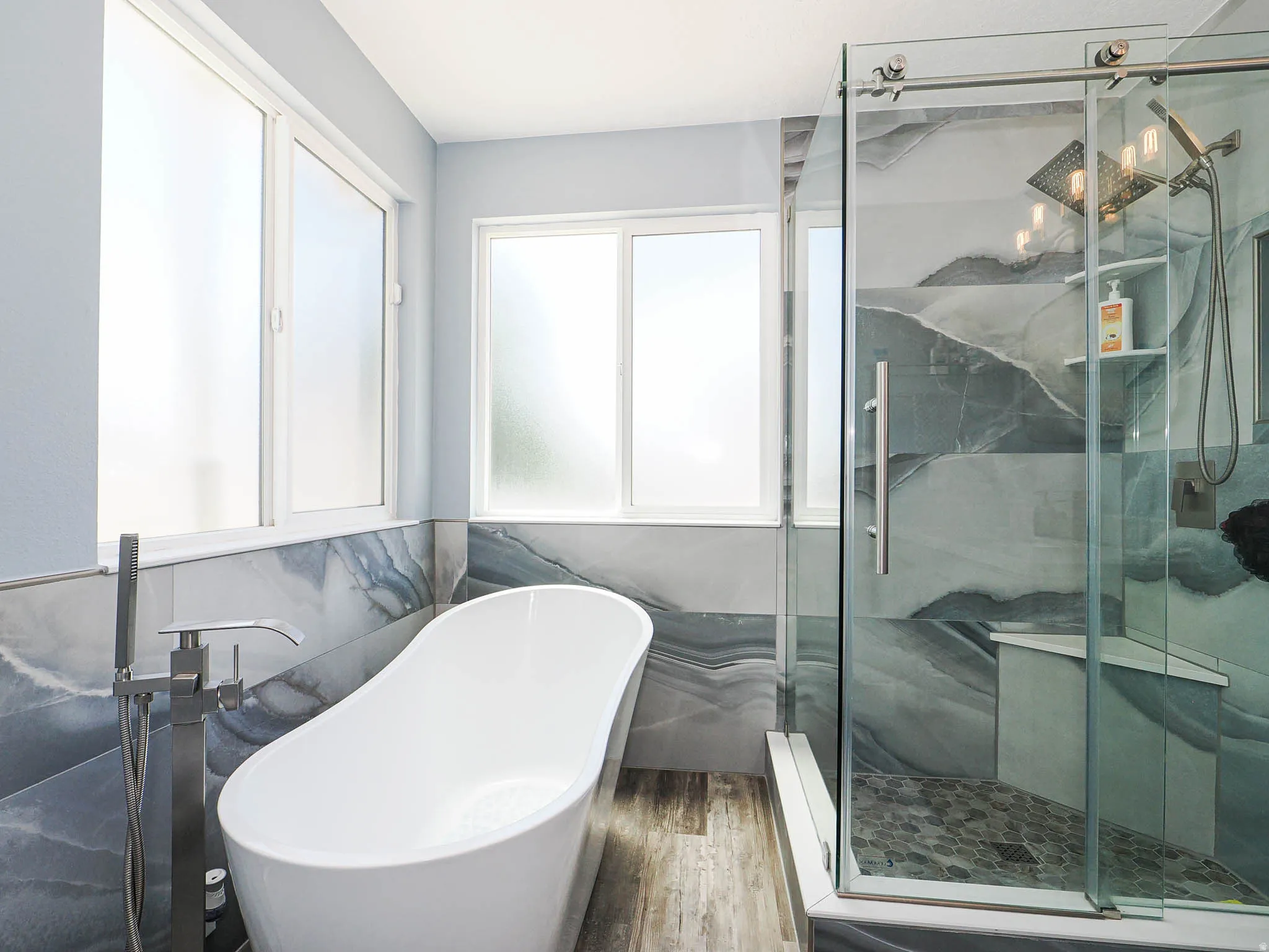 Bathroom featuring a freestanding soaking tub with a floor-mounted faucet, a glass-enclosed shower with tiled walls and a built-in bench, and wood-finish flooring