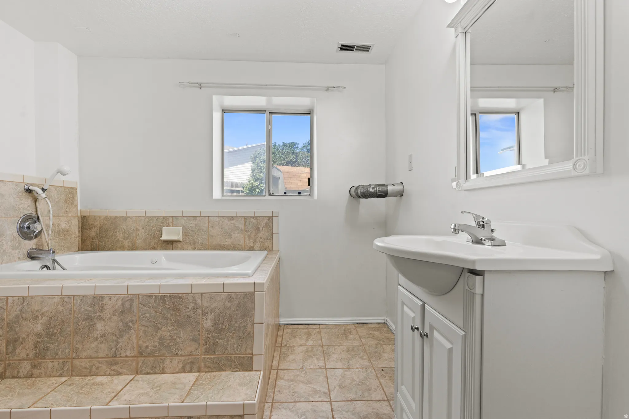 Bathroom featuring a tile-surround drop-in bathtub with a handheld shower fixture, a single vanity with a white basin sink, and a decorative framed mirror
