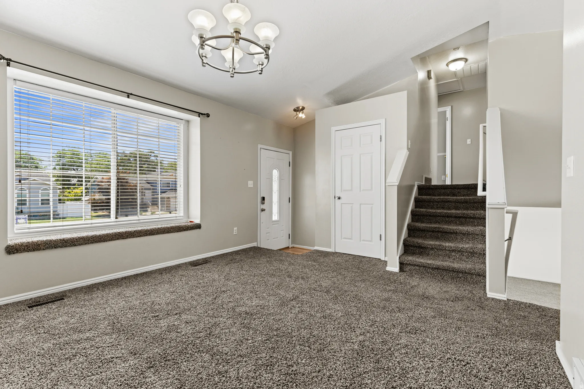 Spacious entry area featuring neutral wall paint, gray carpeting, a large picture window with blinds, a white paneled entry door, and a multi-tiered chandelier