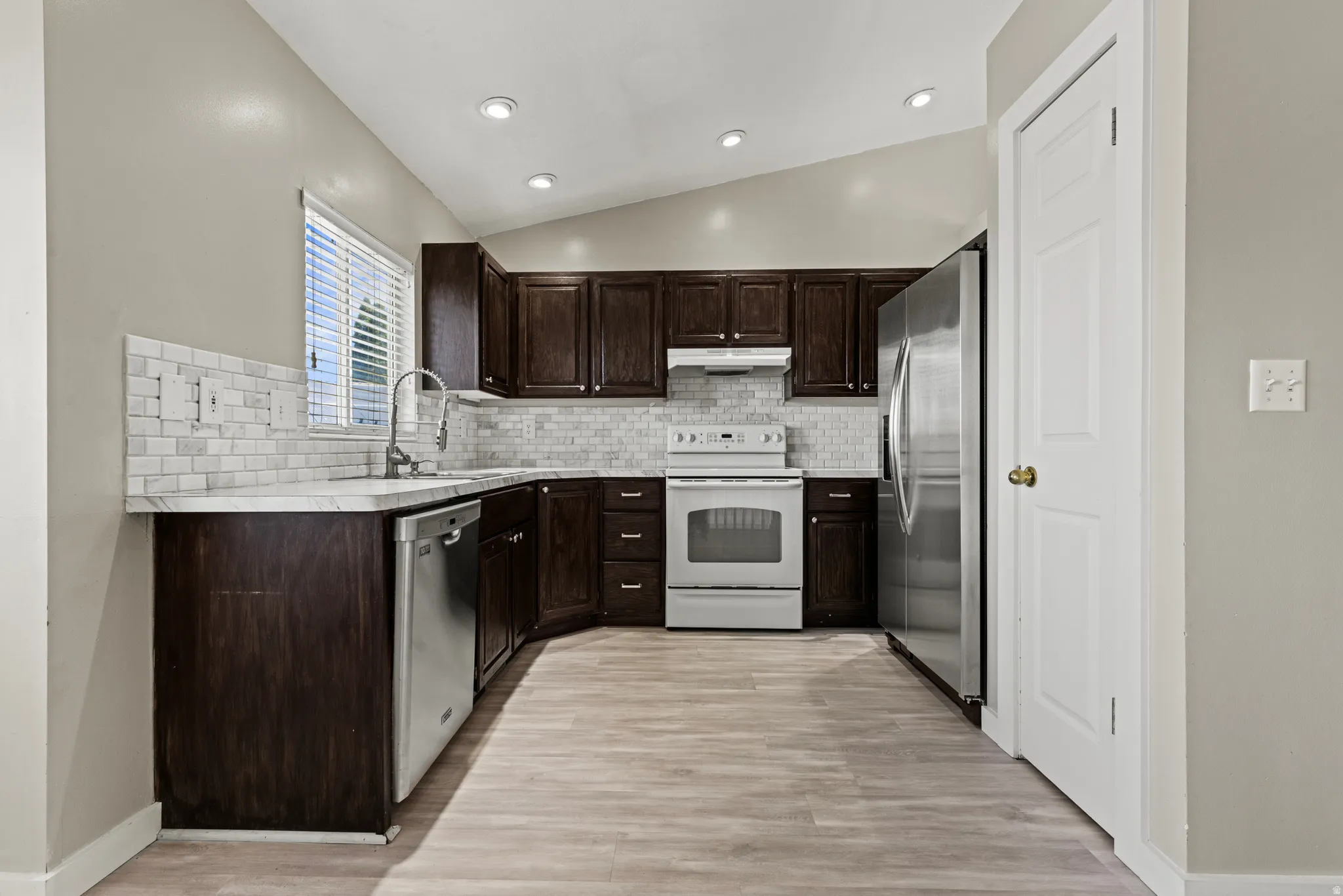 Kitchen featuring dark wood cabinetry, light-toned wood-finish flooring, white subway tile backsplash, stainless steel appliances, and recessed lighting