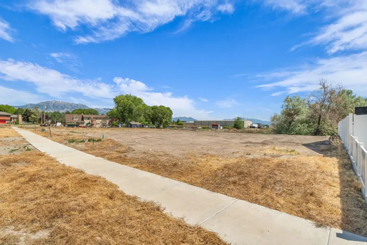 Expansive vacant lot featuring a paved sidewalk, existing perimeter fencing, and distant mountain backdrops