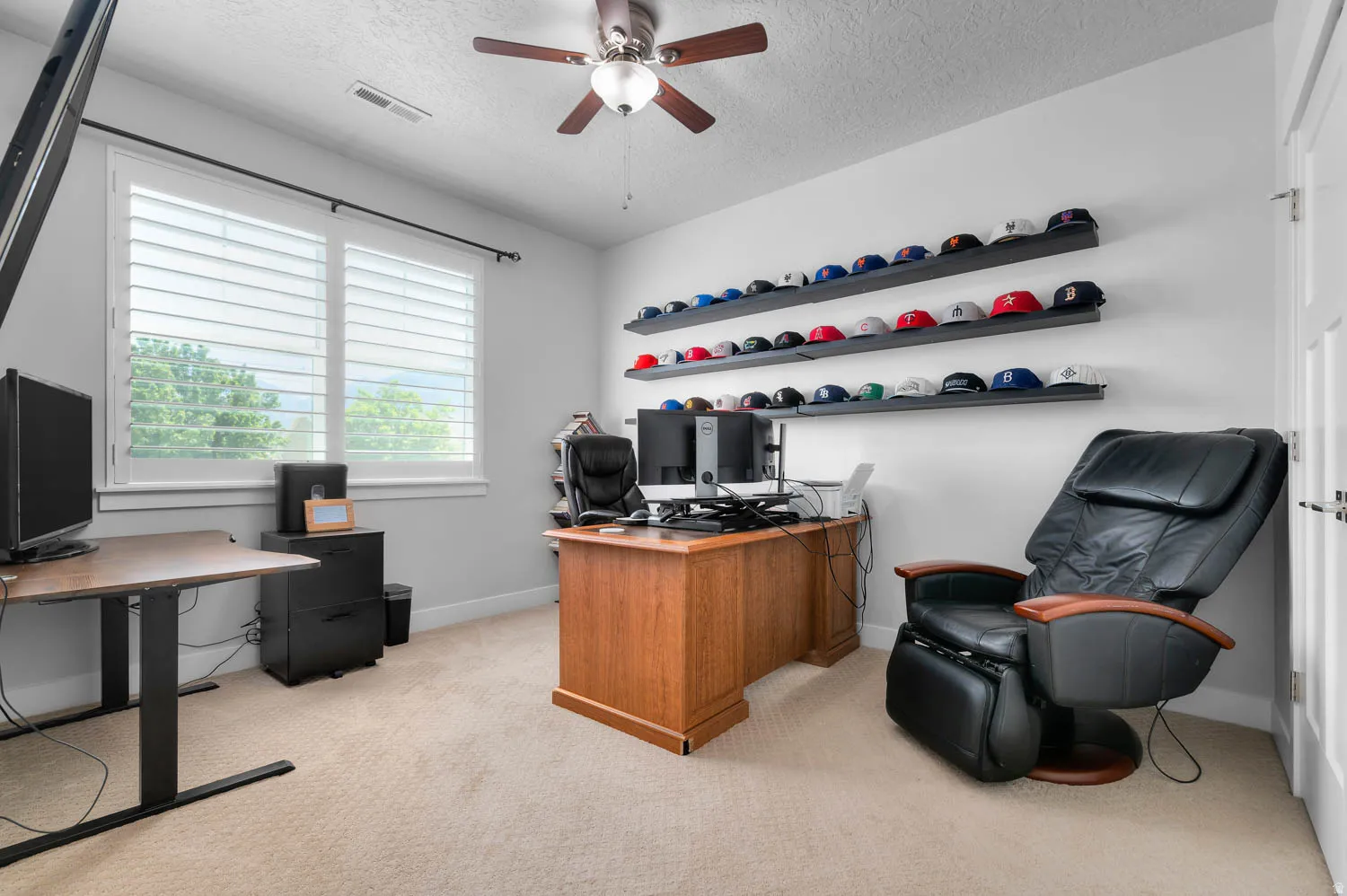Versatile room featuring a large window with plantation shutters, ceiling fan, and light neutral carpet flooring